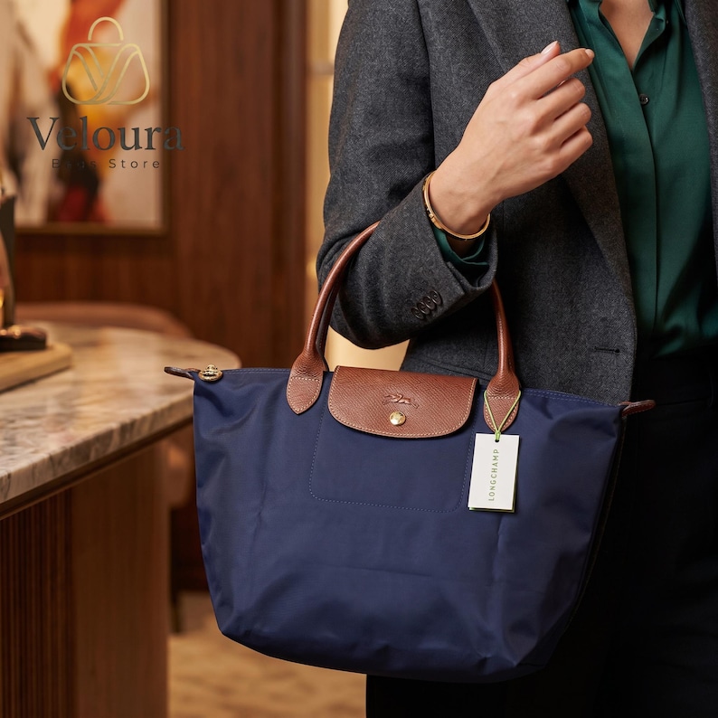 May include: A navy blue tote bag with brown leather handles and a flap closure. The bag has a rectangular shape and a small front pocket. A tag with the brand name is attached to the bag.
