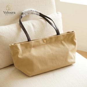 Puede incluir: Un bolso tote de lona beige con asas de cuero marrón oscuro y un cierre de botón a presión de metal dorado en el centro. El bolso tiene una base rectangular y un cuerpo suave y sin estructura. El texto Veloura Bags Store es visible arriba.