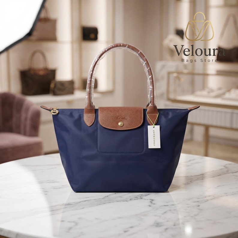 May include: A navy blue tote bag with brown leather accents. The bag has a brown leather flap with a gold-colored button closure and brown leather handles. A white tag with black text hangs from the bag. The bag is on a marble surface.