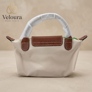 May include: A small, off-white handbag with a brown leather flap and handle accents. The bag features a top handle and a flap closure. The brand name "Veloura Bags Store" is visible in the upper left corner.