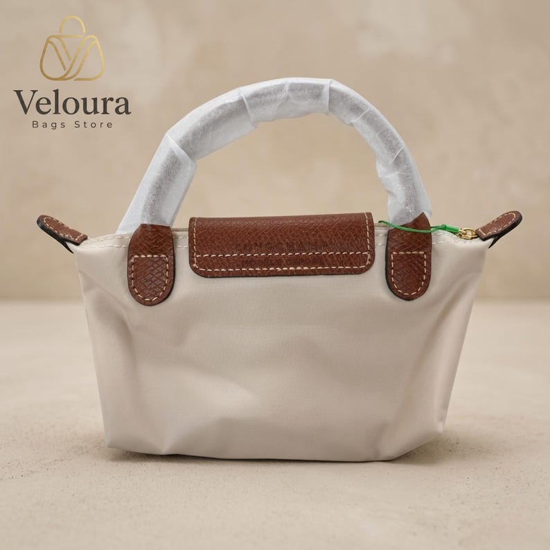 May include: A small, off-white handbag with a brown leather flap and handle accents. The bag features a top handle and a flap closure. The brand name "Veloura Bags Store" is visible in the upper left corner.