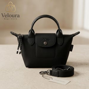 May include: A black handbag with a top handle and a detachable shoulder strap. The bag has a flap closure with a silver button and a small pocket on the front. The bag is made of a smooth material and has a zipper closure. The brand name "Veloura Bags Store" is in the top left corner.
