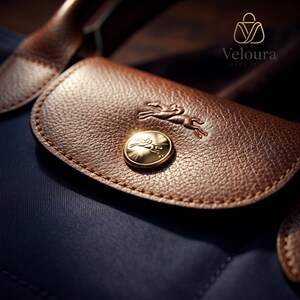 May include: Close-up of a navy blue handbag with a brown leather flap. The flap features a gold emblem of a running horse and a gold button. The bag has a textured leather appearance and a brown leather handle.