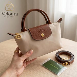 May include: A beige handbag with brown leather accents, including the top flap, handles, and a detachable shoulder strap. The bag features a gold zipper and hardware, with the brand name "Veloura Bags Store" visible in the upper left corner.