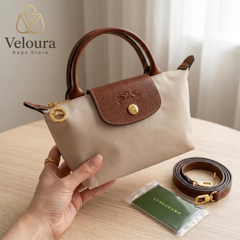 May include: A beige handbag with brown leather accents, including the top flap, handles, and a detachable shoulder strap. The bag features a gold zipper and hardware, with the brand name "Veloura Bags Store" visible in the upper left corner.