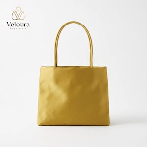 May include: A mustard yellow tote bag with a rectangular body and two matching shoulder straps. The bag features a smooth, solid-colored finish. In the upper left corner, the text Veloura Bags Store is printed with a stylized bag icon above it.