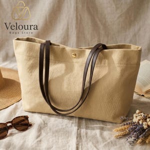 May include: A beige canvas tote bag featuring dark brown leather straps and a single gold-tone metal snap button closure at the top center. The bag has a structured rectangular shape with visible fabric texture and clean stitched seams.