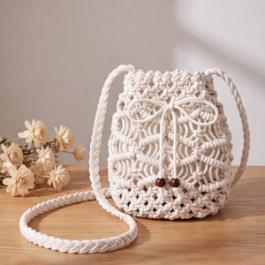 May include: A cream-colored macrame bucket bag featuring an intricate knotted pattern, a braided shoulder strap, and a drawstring closure secured with two dark brown wooden beads at the ends of the cord, creating a textured and functional design.