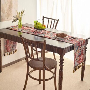 Può includere: Un tavolo da pranzo in legno scuro è decorato con un runner da tavolo fantasia. Un cestino di metallo contiene uva e una pesca. Due sedie di legno sono posizionate attorno al tavolo. Sono presenti anche un vaso con fiori secchi e pane.