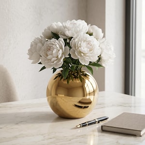 Peut inclure: Un vase sphérique doré à la surface polie et réfléchissante contenant un grand bouquet de pivoines blanches avec des feuilles vertes. Le vase est posé sur une surface en marbre blanc à côté d'un stylo plume noir et doré et d'un carnet gris.