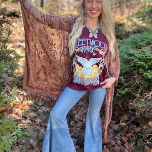 Könnte beinhalten: Eine Frau trägt ein weinrotes Fleetwood Mac Grafik-T-Shirt, hellblaue Schlaghosen und einen braunen, gemusterten Schal mit Fransen. Das T-Shirt zeigt eine Taubengrafik und die Worte "Dreams". Der Hintergrund umfasst Grün und abgefallene Blätter.