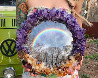 May include: A round mirror surrounded by a frame of amethyst crystals and other stones, reflecting a rainbow and a cloudy sky.