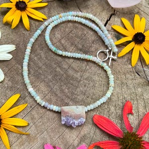 May include: A delicate necklace made with light blue opal beads and a pendant featuring a raw amethyst crystal cluster. The necklace is displayed on a wooden surface with flowers.