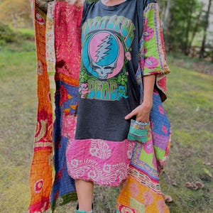 May include: A dark gray t-shirt dress featuring a Grateful Dead graphic with a skull and lightning bolt logo. The dress is layered with a colorful, patchwork-style kimono made of various patterned fabrics in shades of orange, pink, and green.