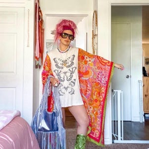Könnte beinhalten: Ein weißes T-Shirt mit schwarzem Schmetterlingsmotiv, ein leuchtend orange-pinkfarbener Kimono mit Blumenmuster, metallisch grüne Cowboystiefel und eine metallisch blaue Handtasche mit Fransen. Das Outfit umfasst eine Sonnenbrille und eine Perlenkette.
