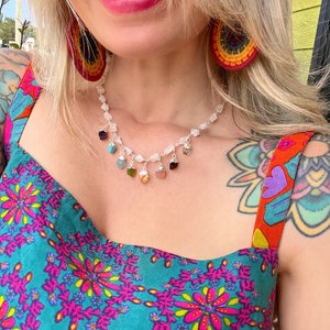 May include: A silver necklace with a white stone chain and colorful gemstone charms. The necklace is worn by a person with a colorful floral pattern shirt and red straps.