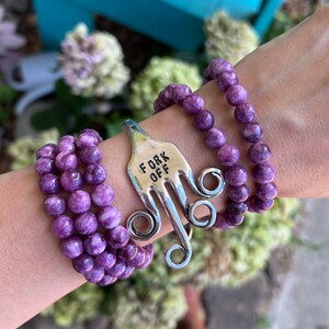May include: A silver fork bracelet with the words "Fork Off" engraved on it. The bracelet is worn on a wrist with two purple beaded bracelets.