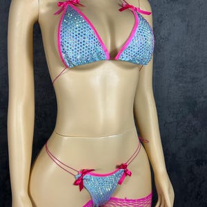 May include: A two-piece bikini set featuring a light blue fabric covered in iridescent rhinestones. The triangle top and thong bottom are trimmed with bright pink fabric and accented with small pink satin bows at the straps and sides.