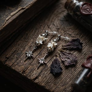May include: A pair of drop earrings featuring a silver-tone post, a small metallic sphere, a white bone-like segment, and a dangling star-shaped charm with a central clear oval crystal. The earrings are arranged with dried dark flower petals.