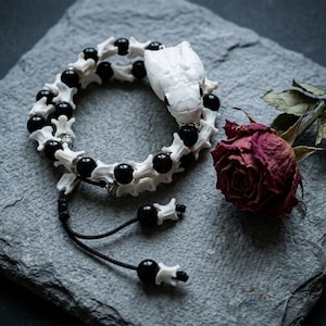 May include: A bracelet featuring a white snake head focal piece, alternating white bone-like vertebrae segments, and smooth black spherical beads. The design includes silver-toned spacers and black cord ends with additional beads and bone segments.