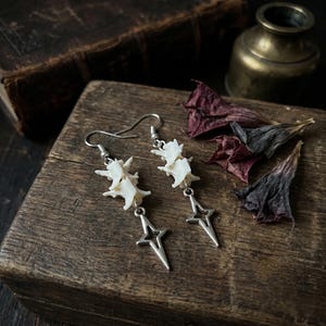 May include: A pair of dangle earrings featuring small, off-white animal vertebrae bones stacked vertically. Each earring is finished with a silver-toned metal star charm at the bottom and a standard silver-toned metal hook for ear attachment.