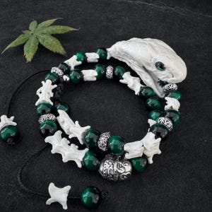 May include: A wrap bracelet featuring a white snake head with green eyes, alternating with dark green beads, white bone-shaped segments, and silver-toned metal spacers. The bracelet includes a black cord and a dangling bone-shaped bead at the end.