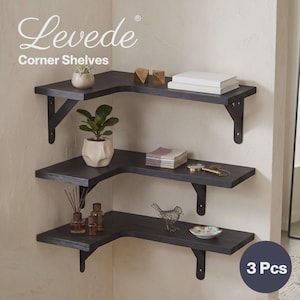 May include: A set of three dark brown L-shaped corner shelves mounted on a wall. Each shelf is supported by two black metal brackets. The shelves hold various decorative items including a small potted plant, books, and small ornaments.