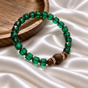 May include: A beaded bracelet featuring faceted emerald green crystal beads arranged in a circular pattern. The design includes two smooth dark brown wooden beads accented by two small gold-toned rings embedded with tiny clear crystals.