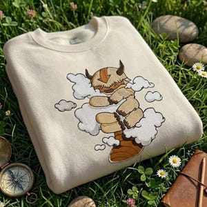May include: A cream-colored crewneck sweatshirt featuring an embroidered design of a tan, furry, six-legged creature with a brown arrow on its head, surrounded by white clouds. The creature has dark brown horns and a striped tail, depicted in a cartoon style.