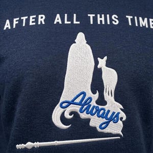 Könnte beinhalten: Ein marineblaues Sweatshirt mit weißem Sticktext, der AFTER ALL THIS TIME lautet. Unter dem Text befindet sich eine weiße Stick-Silhouette einer Kapuzenfigur und eines Hirsches, ergänzt durch das blaue Stickwort Always und einen Zauberstab.