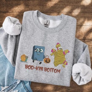 May include: A heather gray crewneck sweatshirt featuring a graphic of a blue square character and a pink starfish character dressed as a mummy, both holding jack-o'-lanterns, with a snail-pumpkin hybrid and the text BOO-KINI BOTTOM printed below.