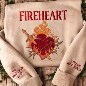 Peut inclure: Un sweat-shirt beige avec le texte brodé en rouge FIREHEART. Le centre présente un cœur rouge brodé entouré de flammes orange, d'une épée et d'épines. Les manches portent le texte rouge TO WHATEVER END et YOU DO NOT YIELD.