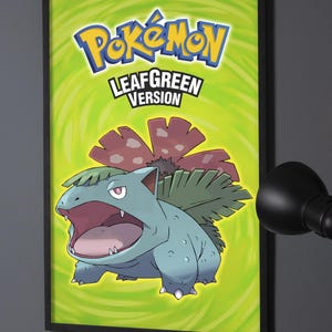 May include: A framed rectangular poster featuring the Pokémon LeafGreen Version logo at the top and a large illustration of the creature Venusaur in the center. The background is a vibrant lime green with a swirling pattern and a black frame.