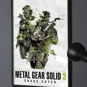 May include: A framed vertical poster featuring a stylised illustration of two characters in tactical gear, one holding a handgun. The text reads TACTICAL ESPIONAGE ACTION METAL GEAR SOLID 3 SNAKE EATER in black and green lettering on a white background.