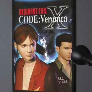 May include: A framed poster featuring the Resident Evil Code: Veronica X video game artwork. The image displays two characters in the foreground, a large letter X, and the text Resident Evil Code: Veronica. A third character is visible in the background.