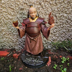 May include: A hand-painted figurine of an elderly character with a topknot, wearing a dark maroon robe with a gold collar. The figure holds a small brown cup in the right hand and a sculpted orange and red flame in the left hand, standing on a base.