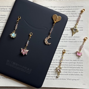 May include: Five gold-toned metal charms with lobster clasps, each featuring small clear beads and distinct pendants including a blue shell, a pink heart with wings, a crescent moon, a star, and a pink butterfly, resting on a black e-reader.
