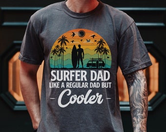 Può includere: T-shirt grigio melange scuro con una grafica in stile vintage di un surfista con una tavola da surf e un furgone. Il testo recita "Surfer Dad Like a Regular Dad But -Cooler-"