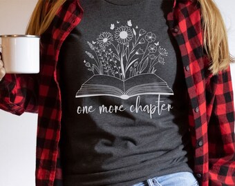 Op de afbeelding: Een donkergrijs heather t-shirt met een witte afbeelding van een open boek met bloemen die eruit groeien. De tekst "one more chapter" staat onder het boek.