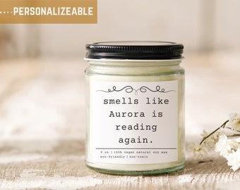 May include: A clear glass jar candle with a black lid and a white label that reads "smells like Aurora is reading again." The label also includes the text "9 oz | 100% vegan natural soy wax eco-friendly | non-toxic".