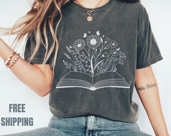 Puede incluir: Una camiseta gris oscuro con un gráfico blanco de un libro abierto con flores y mariposas que salen de él. El texto "Free Shipping" es visible en la parte inferior de la imagen.