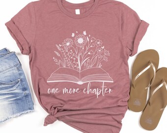 May include: A heather mauve t-shirt featuring a floral book design and the words "one more chapter" in white. The shirt is styled with denim shorts and brown flip-flops, creating a casual, book-themed outfit.