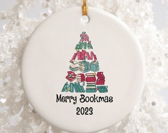 Könnte beinhalten: Weißer Keramik-Weihnachtsbaumschmuck mit einem Weihnachtsbaum aus gestapelten Büchern. Der Baum ist mit roten und grünen Kugeln geschmückt. Der Text "Merry Bookmas 2023" ist unter dem Baum gedruckt.