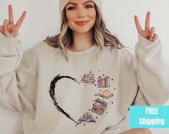 May include: A light beige sweatshirt with a black heart design and a stack of colorful books inside the heart. The books are arranged in a stack with the top book open. The sweatshirt has a crew neck and long sleeves.