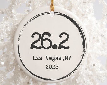 Puede incluir: Adorno de cerámica blanca con un círculo negro y el texto "26.2 Las Vegas, NV 2023".