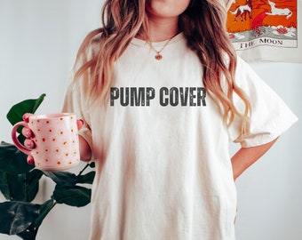 Può includere: Una donna indossa una maglietta bianca oversize con la scritta "PUMP COVER" stampata su di essa. Sta tenendo una tazza rosa a pois. La maglietta è avorio.