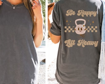 May include: A dark gray t-shirt with the text "Be Happy Lift Heavy" in a retro font. A cartoon kettlebell with a smiling face and a flower is in the center of the text.