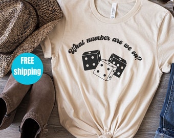 Könnte beinhalten: Ein beiges T-Shirt mit dem Text "What number are we on?" und drei schwarzen Würfeln, die auf die Vorderseite gedruckt sind. Das Shirt liegt auf einer Holzoberfläche mit einem Cowboyhut, Stiefeln und Jeans.