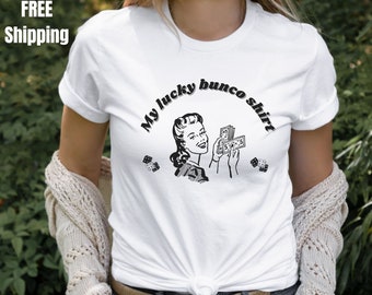 Könnte beinhalten: Weißes T-Shirt mit einer Schwarzweißillustration einer Frau, die Geld und Würfel hält. Der Text "My lucky bunco shirt" ist in einem schwarzen Bogen über der Illustration gedruckt.