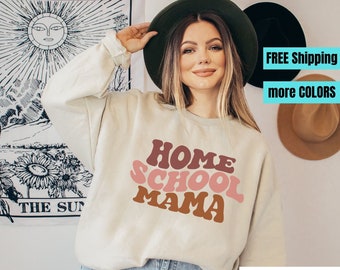 Puede incluir: Una sudadera color arena con el texto "HOME SCHOOL MAMA" en tonos marrones y rosas. La sudadera tiene mangas largas y cuello redondo. La dirección del sitio web www.HollandDriveStudios.com es visible en la parte inferior.
